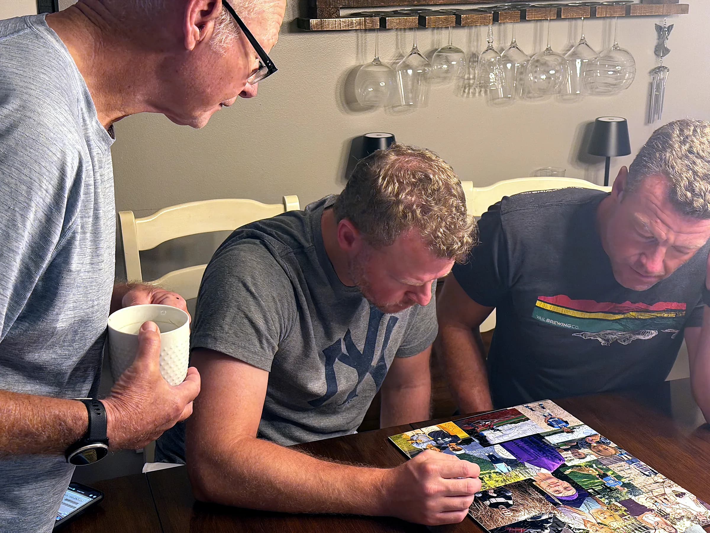 Three people leaning over a collage puzzle in progress at a dining table